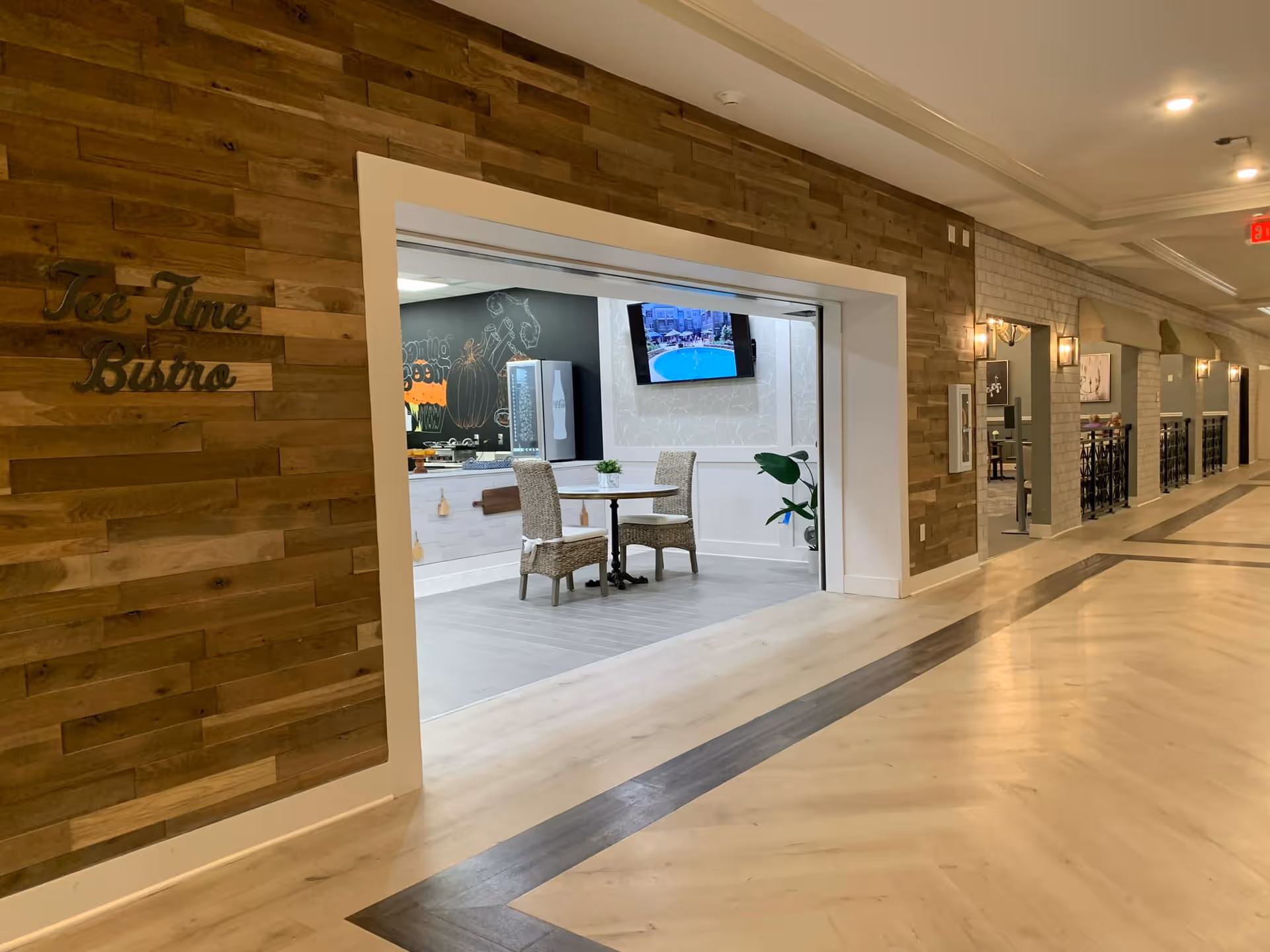 Interior view of a hallway in a senior living facility with light wood flooring and a wooden accent wall. A large open doorway reveals a small dining area with a round table and two wicker chairs. A flat-screen TV is mounted on the wall inside the dining area, and a plant is placed near the corner. The wooden accent wall has the words 'Tea Time Bistro' mounted on it.