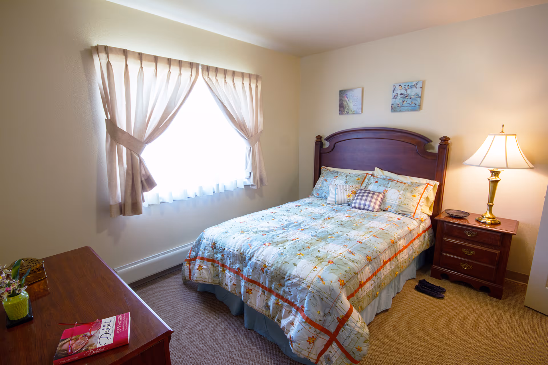 A cozy bedroom with a double bed featuring a floral patterned comforter and multiple pillows. There is a wooden nightstand with a brass lamp and a small dish, and a wooden dresser with a small plant and a book on top. The room has beige walls, a window with beige curtains letting in natural light, and two framed pictures hanging above the bed.