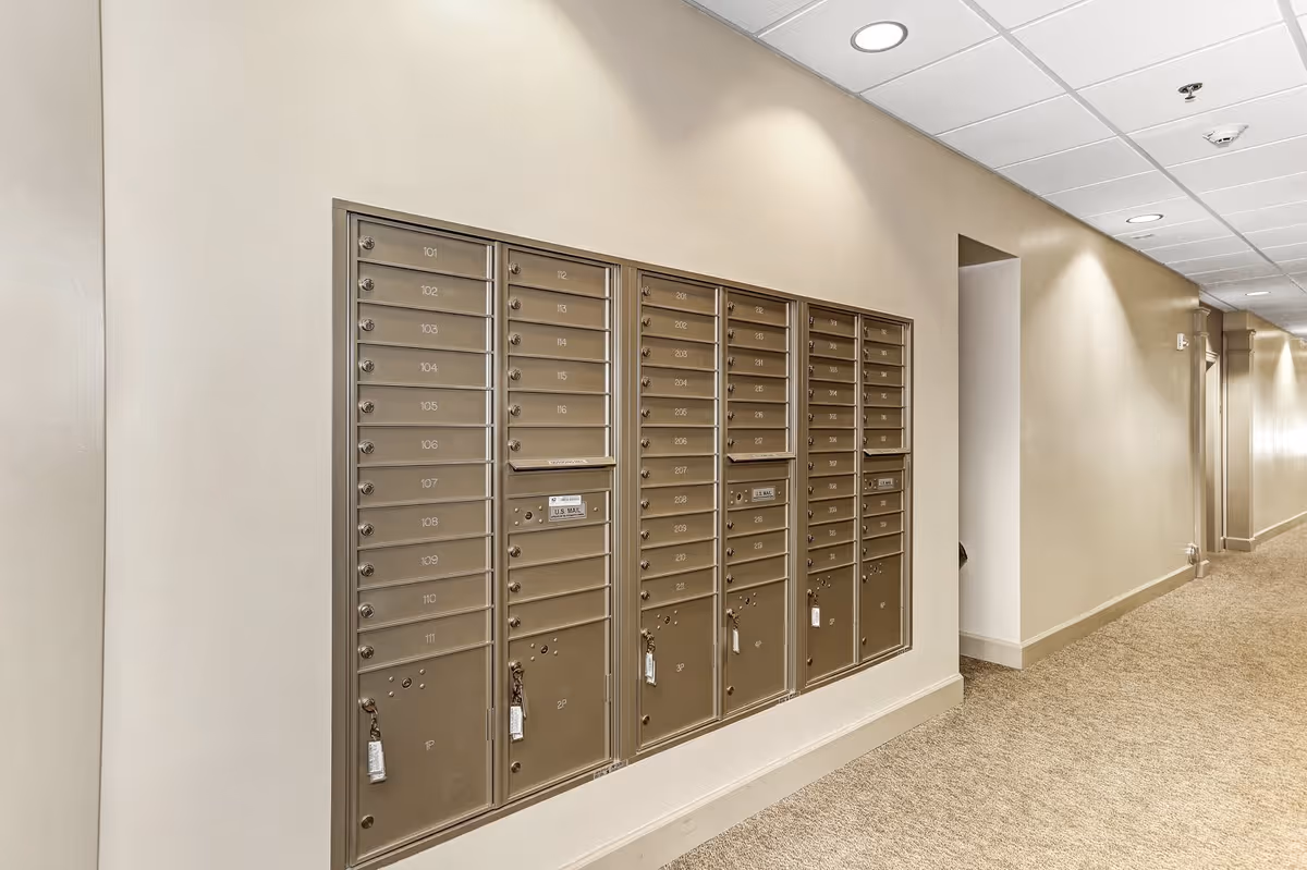 Indoor hallway with a set of metal mailboxes mounted on the wall, each labeled with numbers and some with locks. The hallway has beige walls, carpeted floor, and recessed ceiling lights.