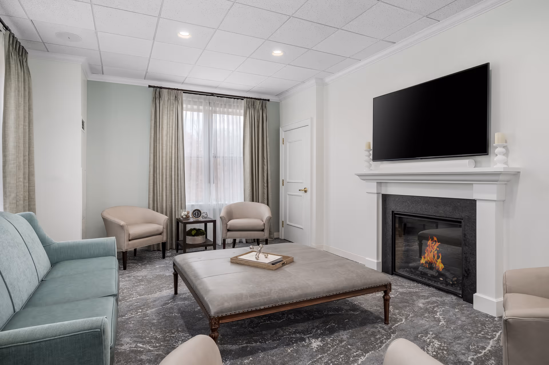 A cozy living room with a light blue sofa, two beige armchairs, a large square ottoman in the center with a tray on top, a fireplace with a fire burning, and a large flat-screen TV mounted above the fireplace. The room has light-colored walls, a window with sheer and beige curtains, and a gray patterned carpet.