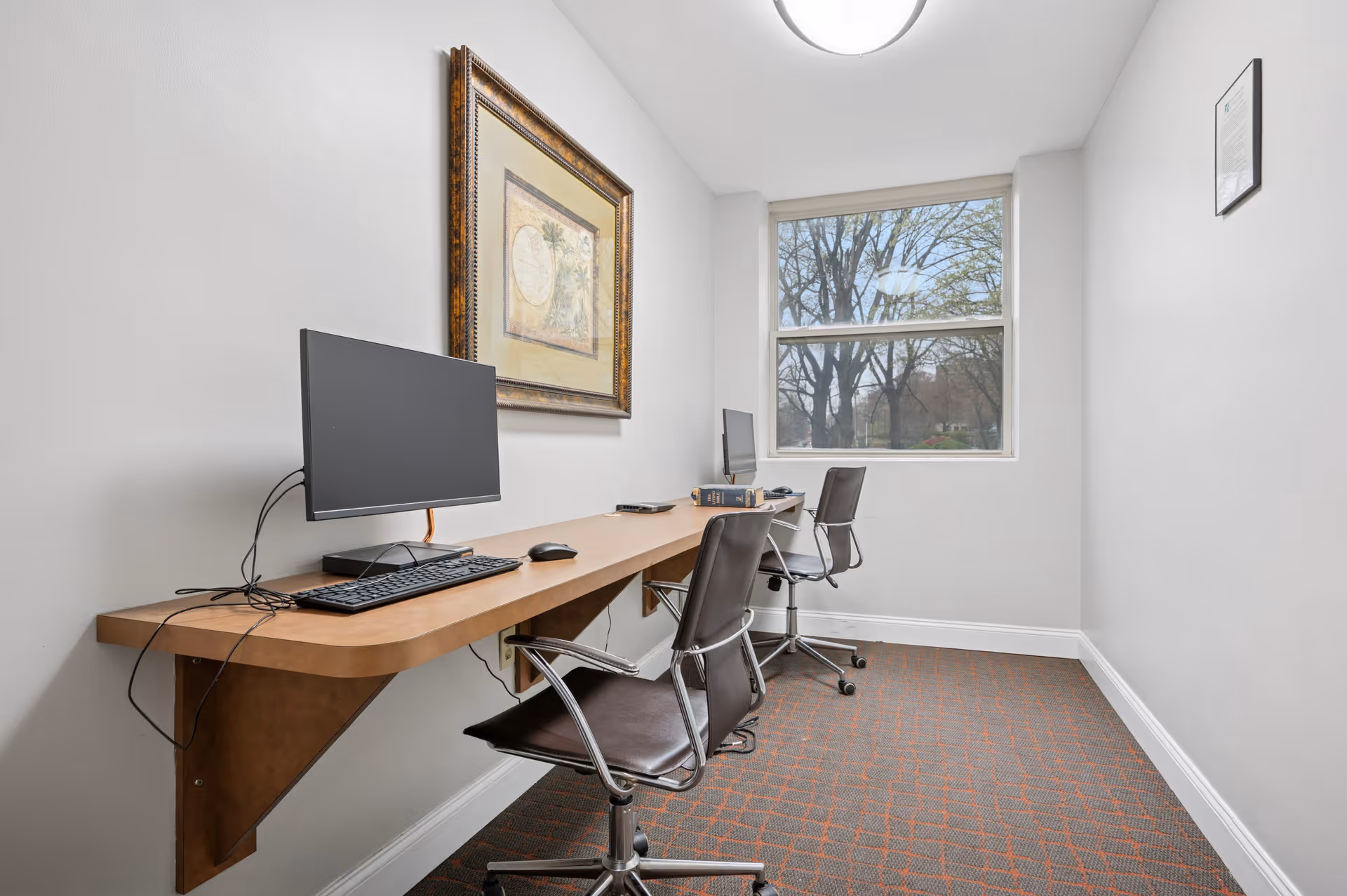 A small office or computer room with two workstations along one wall. Each workstation has a computer monitor, keyboard, and mouse on a long wooden desk mounted to the wall. Two black office chairs with wheels are positioned at the desks. A framed artwork hangs on the wall above the first workstation. A large window at the end of the room shows trees outside. The room has white walls and a carpeted floor with a red and gray pattern.