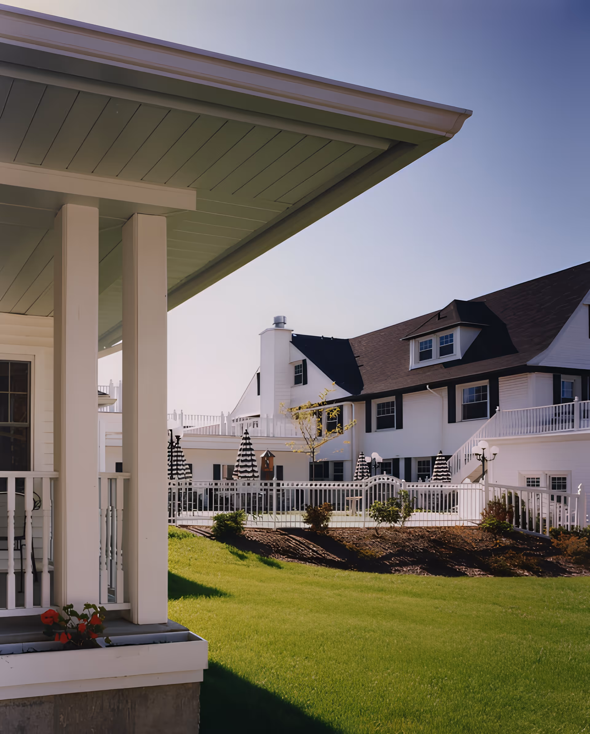 White multi-story residential building with porches, a fenced courtyard and striped umbrellas, viewed from a covered porch over a green lawn.