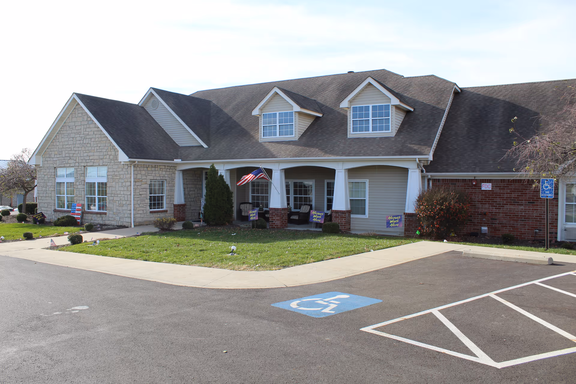 Exterior view of a single-story senior living facility building with a combination of stone and brick facade, a covered porch with white columns, an American flag, and a handicapped parking space in front.