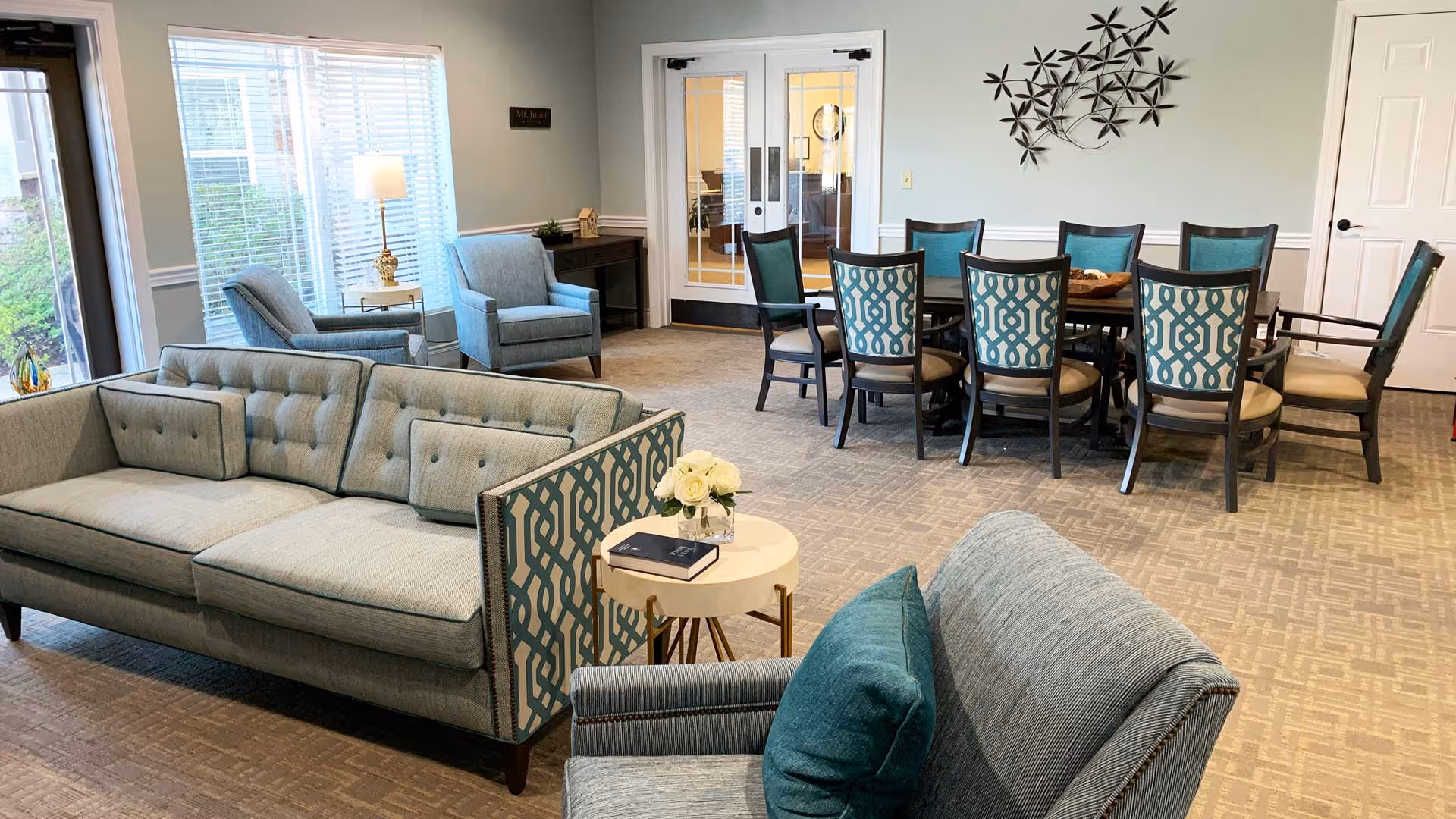 Bright communal living area with teal sofas and armchairs, a round dining table with patterned chairs, and wall decor.