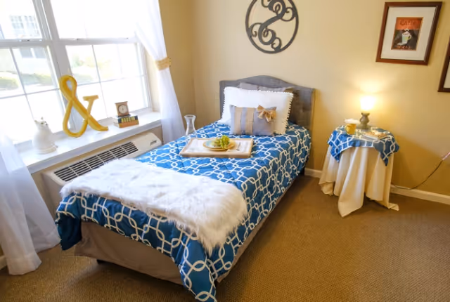 A cozy bedroom with a single bed covered in a blue and white patterned bedspread, a white furry throw at the foot of the bed, and decorative pillows. A tray with grapes and a glass is placed on the bed. Next to the bed is a small round table with a blue and white tablecloth, a lamp, and a cup. The room has beige walls, carpeted floor, a window with white curtains, and decorative wall art above the bed.