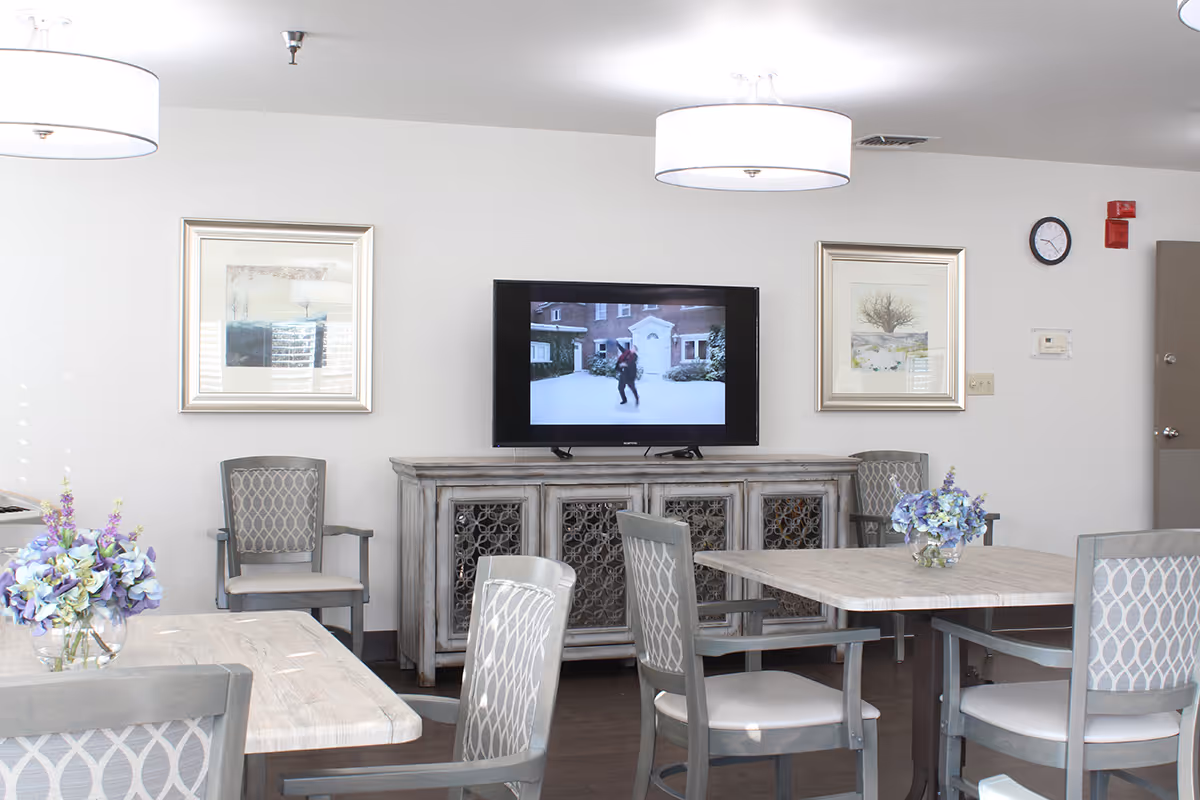 A bright common area with light-colored walls and dark wood flooring featuring a flat-screen TV mounted on the wall above a rustic wooden cabinet. There are several gray chairs with patterned backs around light wood tables, each table decorated with a vase of purple and white flowers. Two framed pictures hang on the wall on either side of the TV, and two modern ceiling lights illuminate the room.