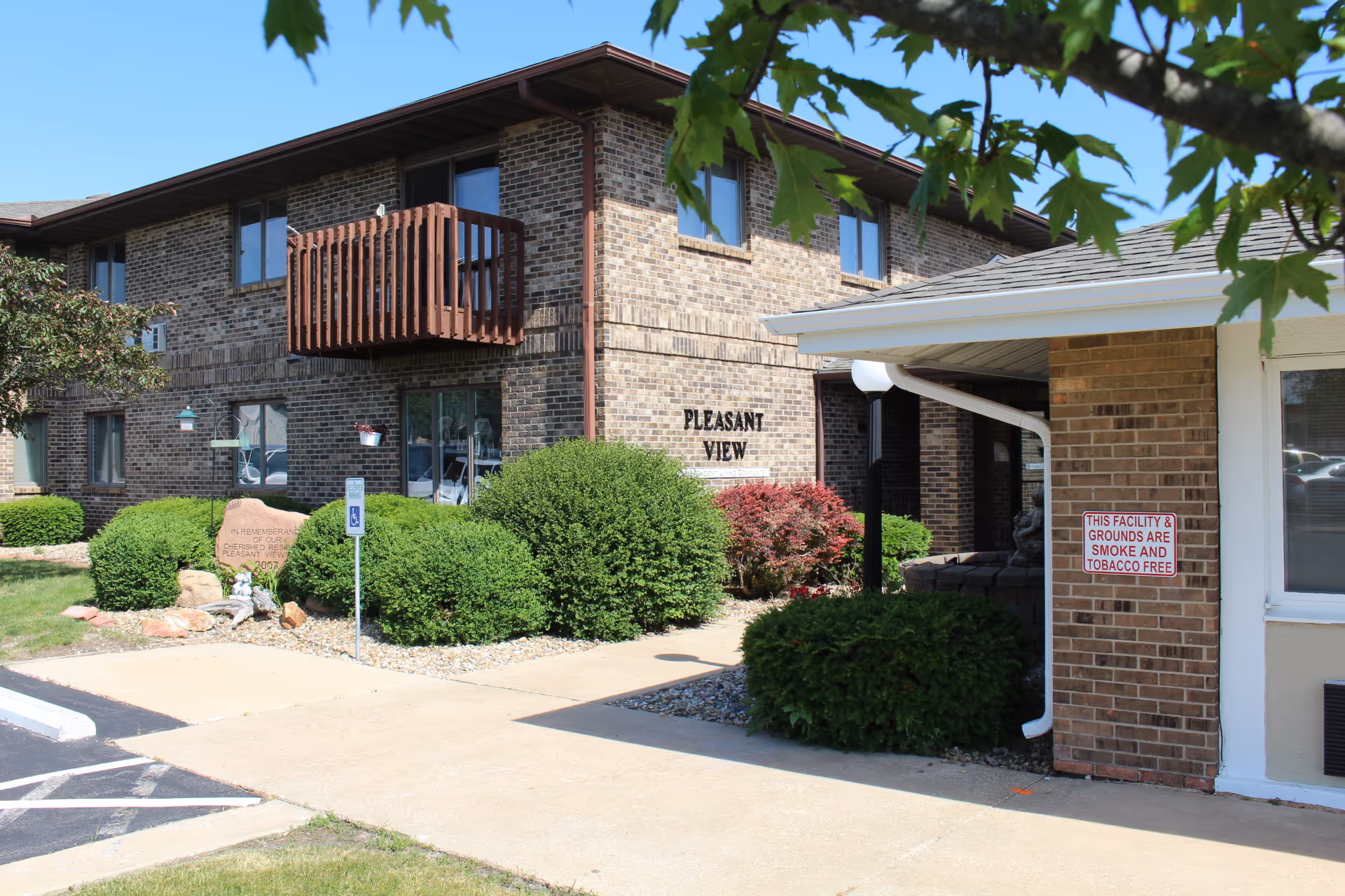 Exterior view of a two-story brick building with a small wooden balcony and several windows. The building has a sign that reads 'PLEASANT VIEW' and is surrounded by green bushes and a sidewalk. A sign on a brick wall near the entrance states that the facility and grounds are smoke and tobacco free. The sky is clear and blue, and some tree leaves are visible in the foreground.