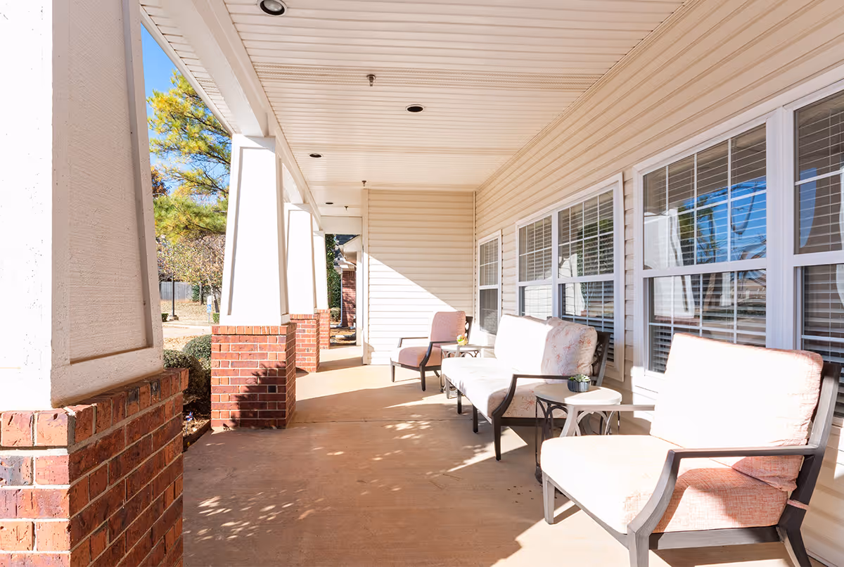 A sunny covered porch area with cushioned chairs and a small table, adjacent to a building with multiple windows and beige siding. Brick pillars support the porch roof, and trees and shrubs are visible in the background.