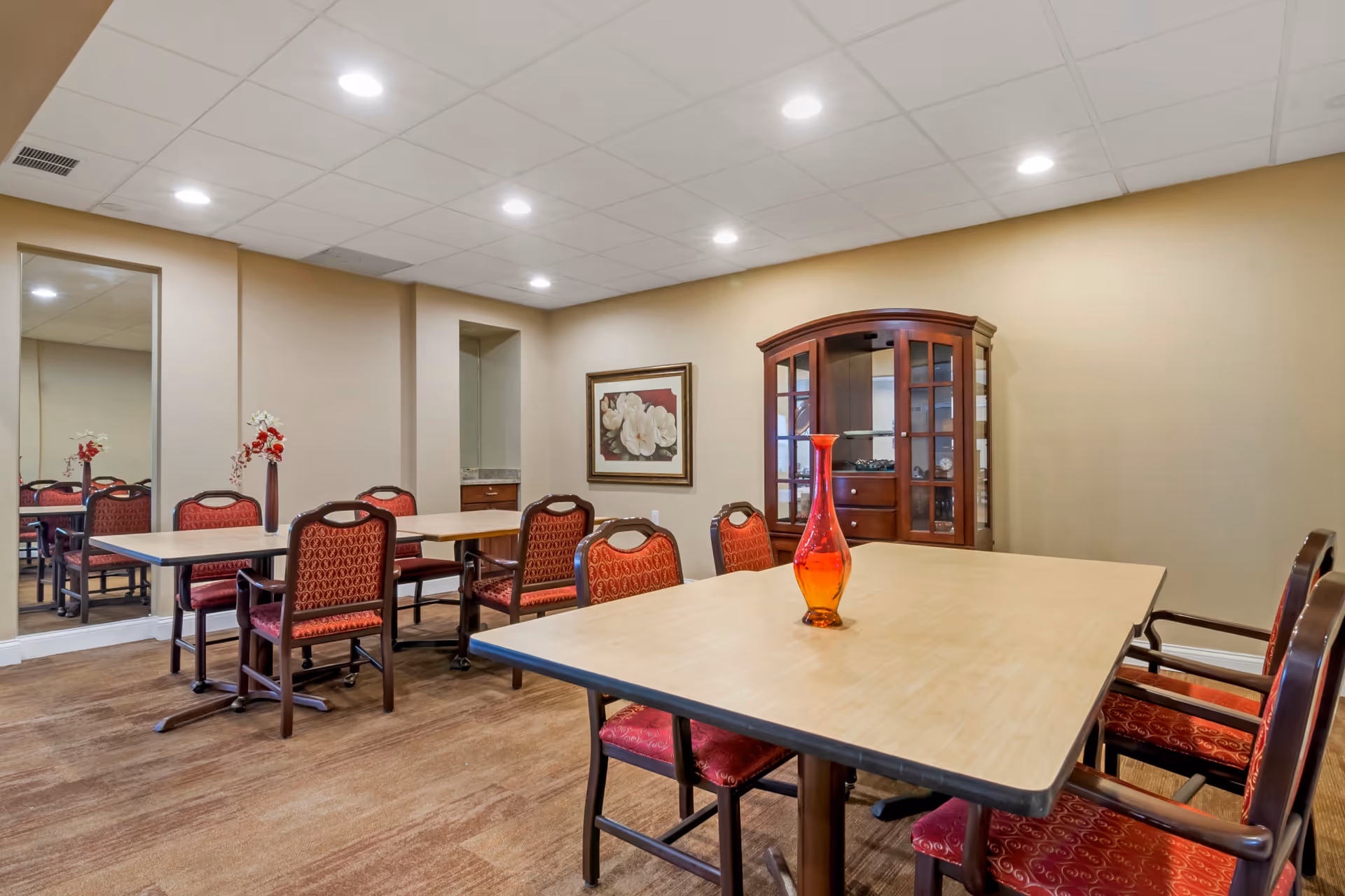 A senior living facility dining room with beige walls and ceiling lights. The room contains two rectangular tables with red cushioned chairs around them. A red and orange decorative vase is placed on the table in the foreground. There is a wooden cabinet with glass doors against the wall and a framed floral painting hanging beside it. A large mirror is mounted on the left wall reflecting part of the room.