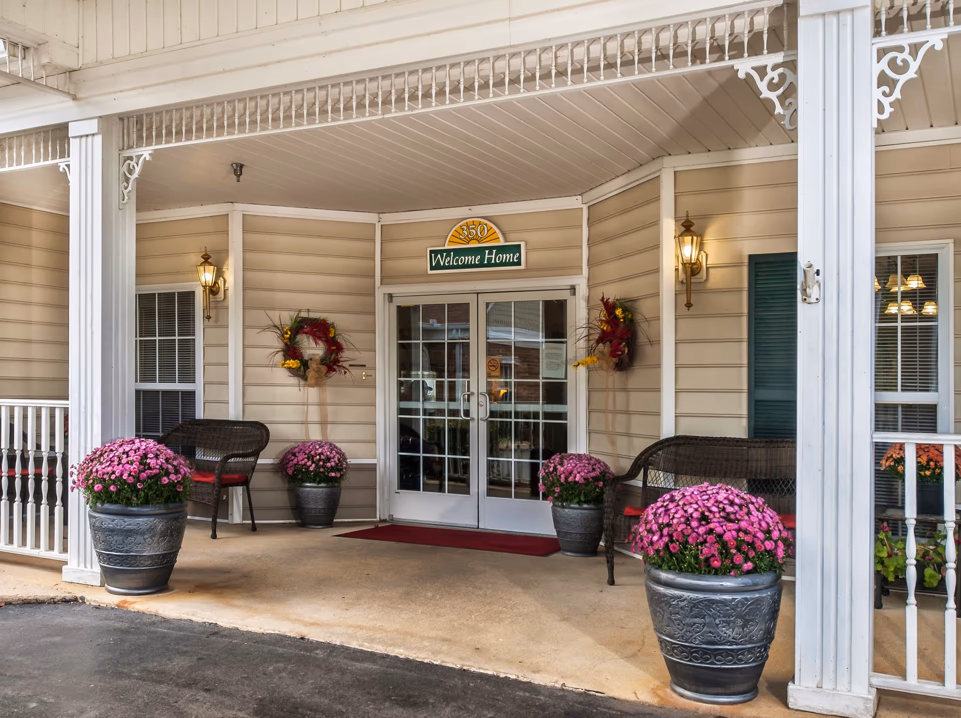 Entrance to a senior living facility with double glass doors under a covered porch. The beige siding walls are decorated with two wreaths and two wall-mounted lantern lights. There are four large pots with pink flowers placed symmetrically around the entrance. Two wicker benches with red cushions are positioned on either side of the doors. Above the doors is a sign that reads '350 Welcome Home'.