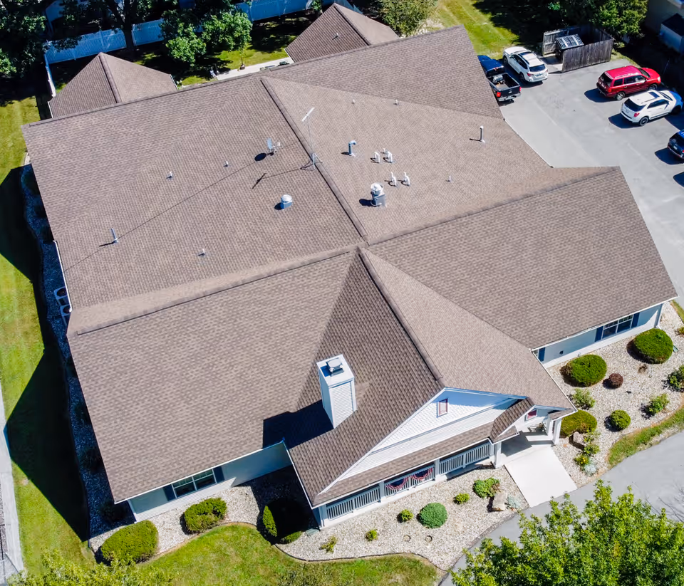 Aerial view of a single-story building with a brown shingled roof, surrounded by green lawns, bushes, and a parking lot with several cars.