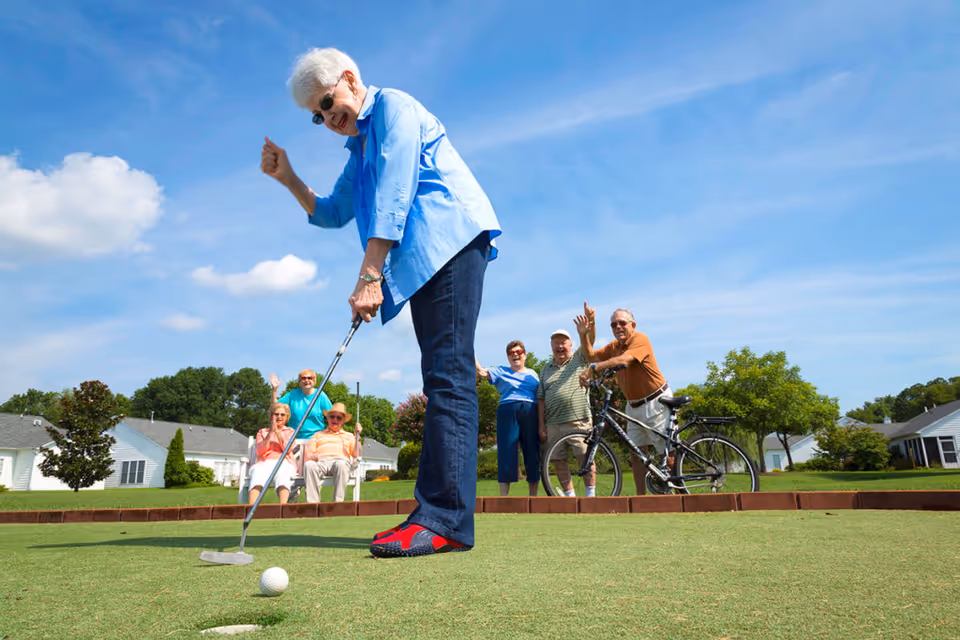 An elderly woman wearing sunglasses and a blue shirt is putting a golf ball on a green while five other elderly people watch and wave in the background. They are outdoors on a sunny day with houses and trees visible behind them.