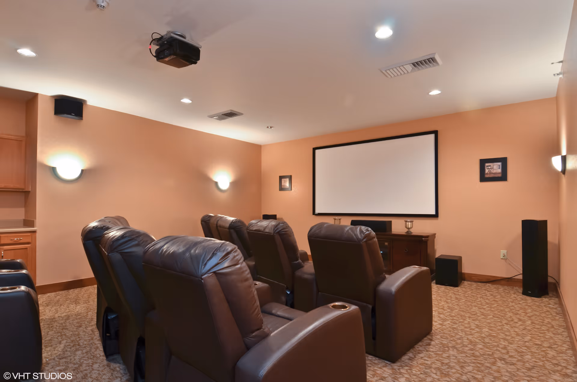 A small home theater room with six brown leather recliner chairs arranged in two rows facing a large blank projection screen mounted on a peach-colored wall. The room has carpeted flooring, wall-mounted lights, a ceiling projector, and speakers positioned around the room.