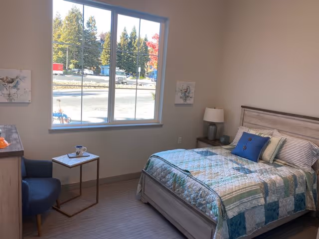 A bright bedroom with a large window showing trees outside. The room features a bed with a patchwork quilt and several pillows, a bedside table with a lamp, a small side table with a cup and saucer, a blue upholstered chair, and a dresser with a decorative item on top. Two small paintings hang on the walls.