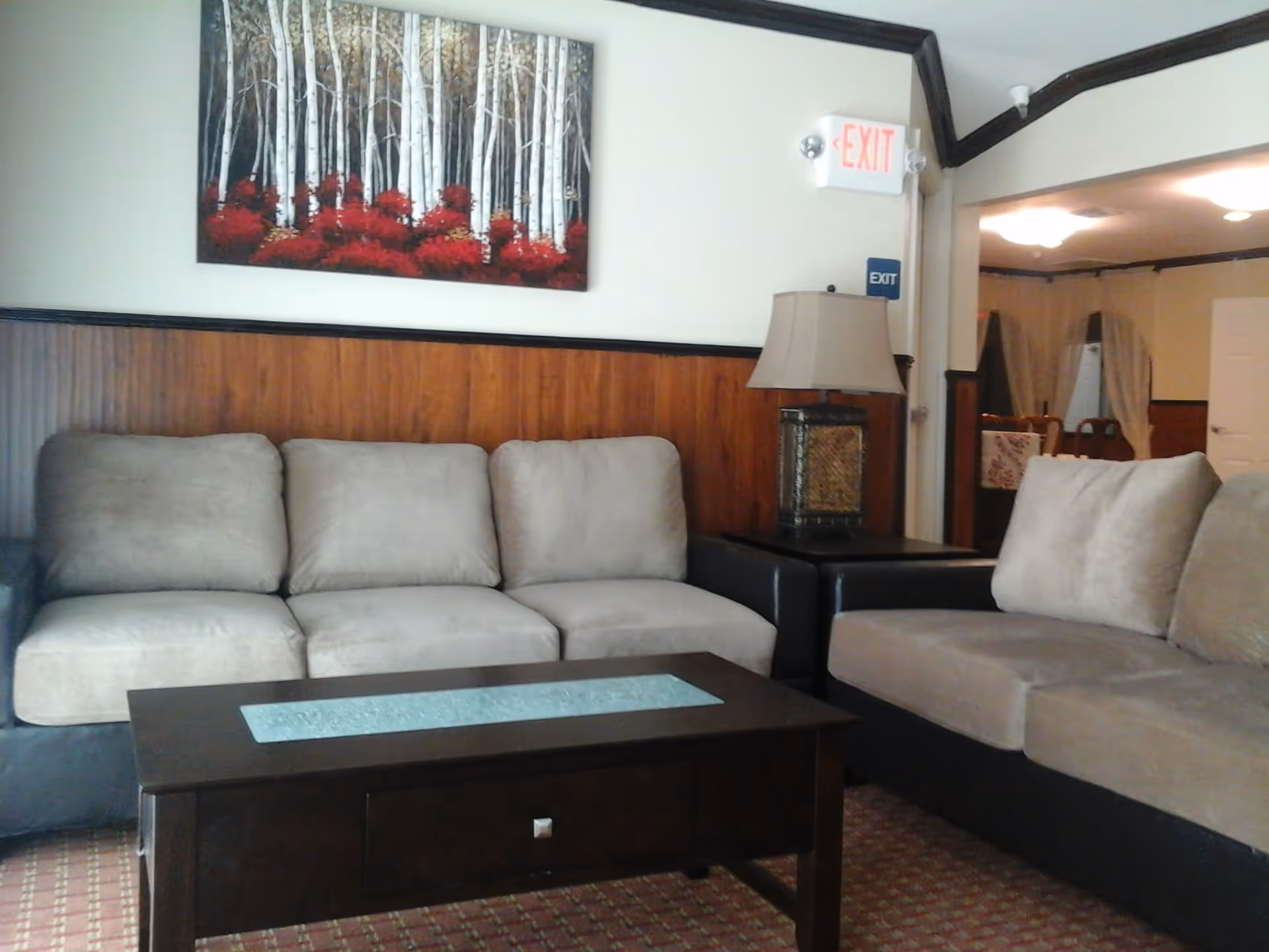 A cozy living room area with two beige sofas facing a dark wooden coffee table. A decorative lamp sits on a side table between the sofas. On the wall above the sofa is a painting of white birch trees with red foliage at the base. An exit sign is visible above the lamp, and a dining area with chairs and a table can be seen in the background.