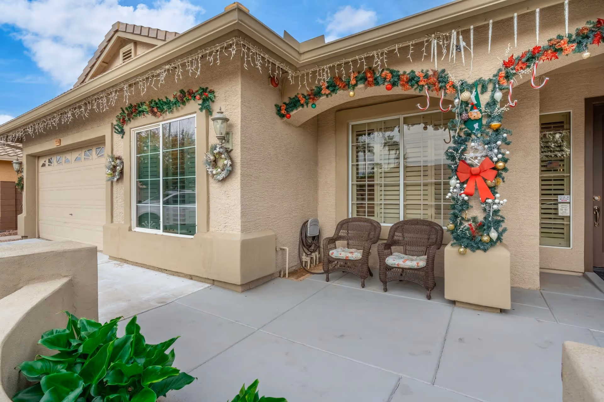 Front entrance of a beige stucco building decorated for Christmas with garlands, wreaths, string lights, two wicker chairs on the porch, and an attached garage.