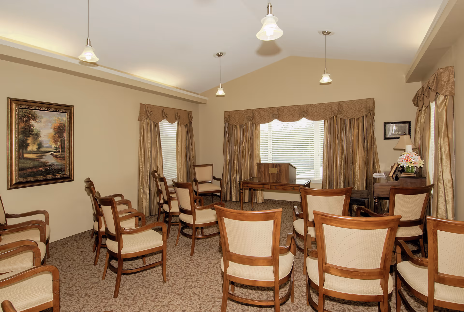 A small meeting or gathering room with several wooden chairs arranged in rows facing a wooden podium and a table near a large window with beige curtains. The room has beige walls, carpeted floor, framed artwork on the wall, and pendant lights hanging from the ceiling.