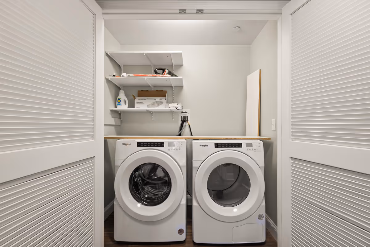 A laundry room with a front-loading washing machine and dryer side by side under a wooden countertop. Above them are three white shelves holding laundry supplies and other items. The room has white louvered doors partially open and light gray walls.