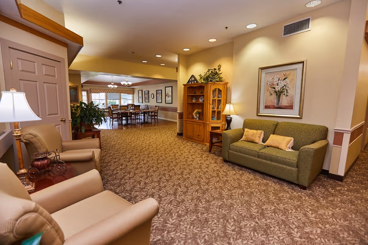 A cozy senior living facility common area with beige armchairs and a green sofa with two yellow pillows. A wooden cabinet with decorative items and a table lamp is against the wall, which also features a framed floral painting. In the background, there is a dining area with tables and chairs near large windows letting in natural light.