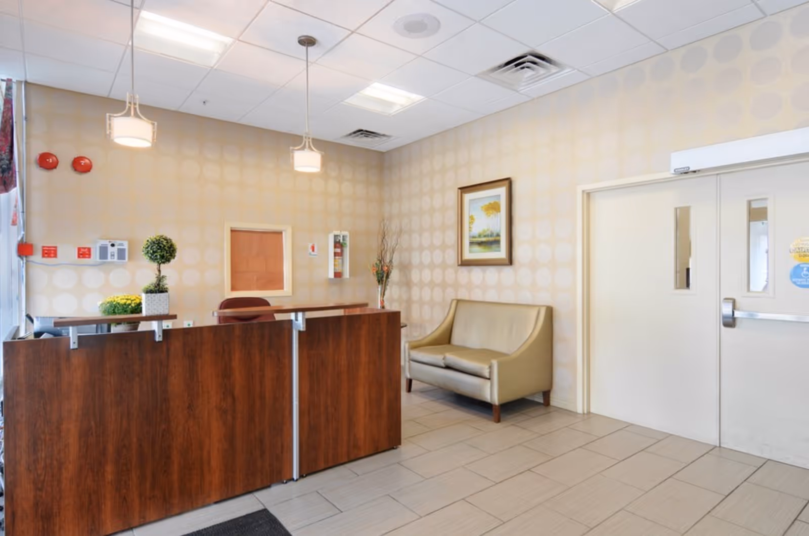Reception area with a wooden front desk, a beige loveseat, a framed painting on the wall, two hanging ceiling lights, and a tiled floor. There are double doors to the right and some decorative plants on the desk and in the corner.