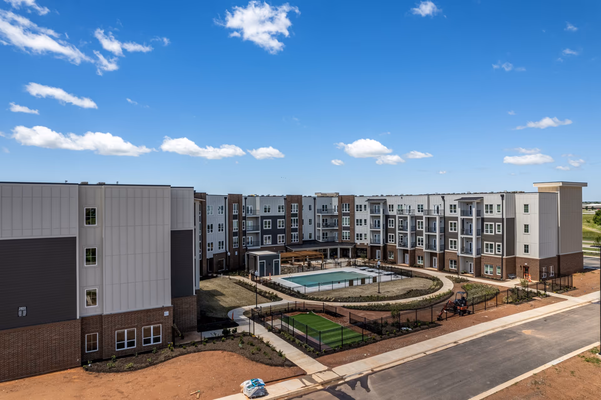 Exterior view of a modern four-story senior living facility with a central courtyard featuring a swimming pool, walking paths, and landscaped areas under a blue sky with scattered clouds.