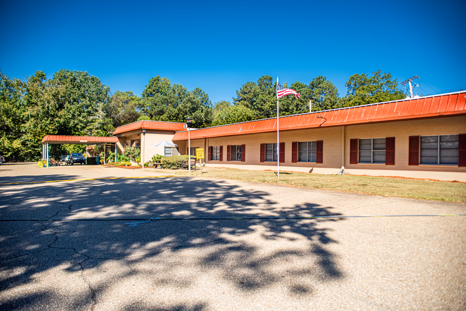 Exterior view of a single-story building with a red roof and beige walls, featuring several windows with red shutters. An American flag is flying on a flagpole in front of the building. There is a covered entrance area on the left side with a car parked underneath. Trees and greenery surround the building under a clear blue sky.
