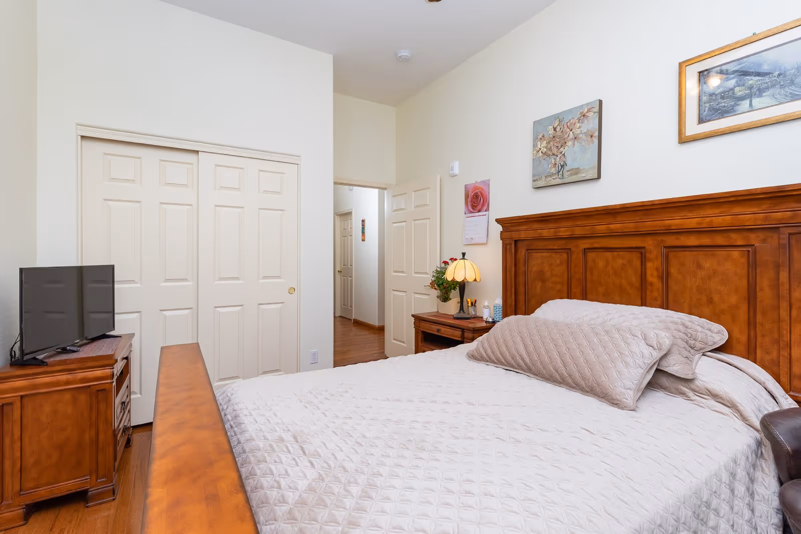 A bedroom with a large wooden bed frame and a beige quilted bedspread and pillows. There is a wooden nightstand with a lamp, a potted plant, and some small items. On the wall above the bed are two framed pictures. A wooden dresser with a flat-screen TV is positioned near a set of white sliding closet doors. The room has light-colored walls and wooden flooring.