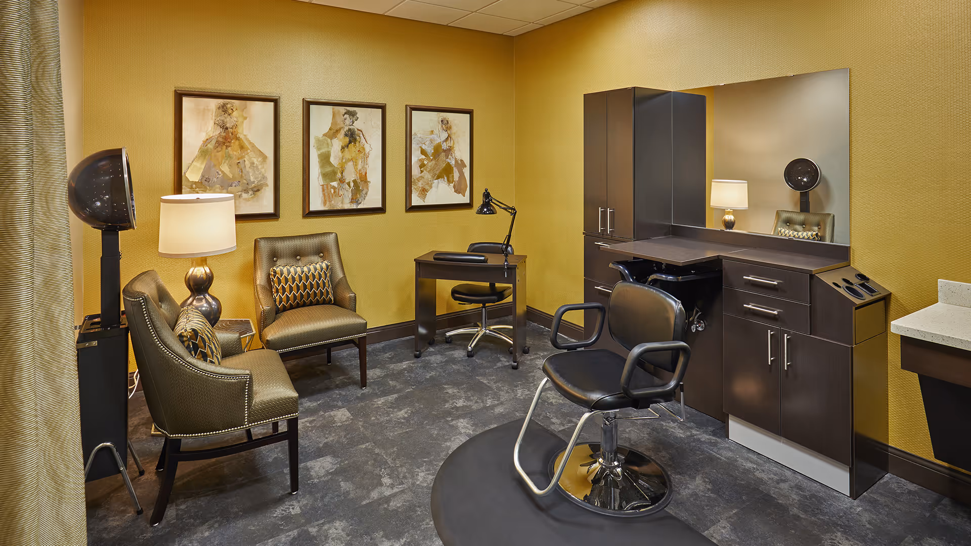 A well-lit salon room with mustard yellow walls featuring three framed abstract paintings. The room contains a black salon chair on a circular mat in front of a large mirror and dark wood cabinetry. There are two upholstered chairs with patterned cushions, a small desk with a black swivel chair, a floor lamp, and a vintage-style hair dryer.