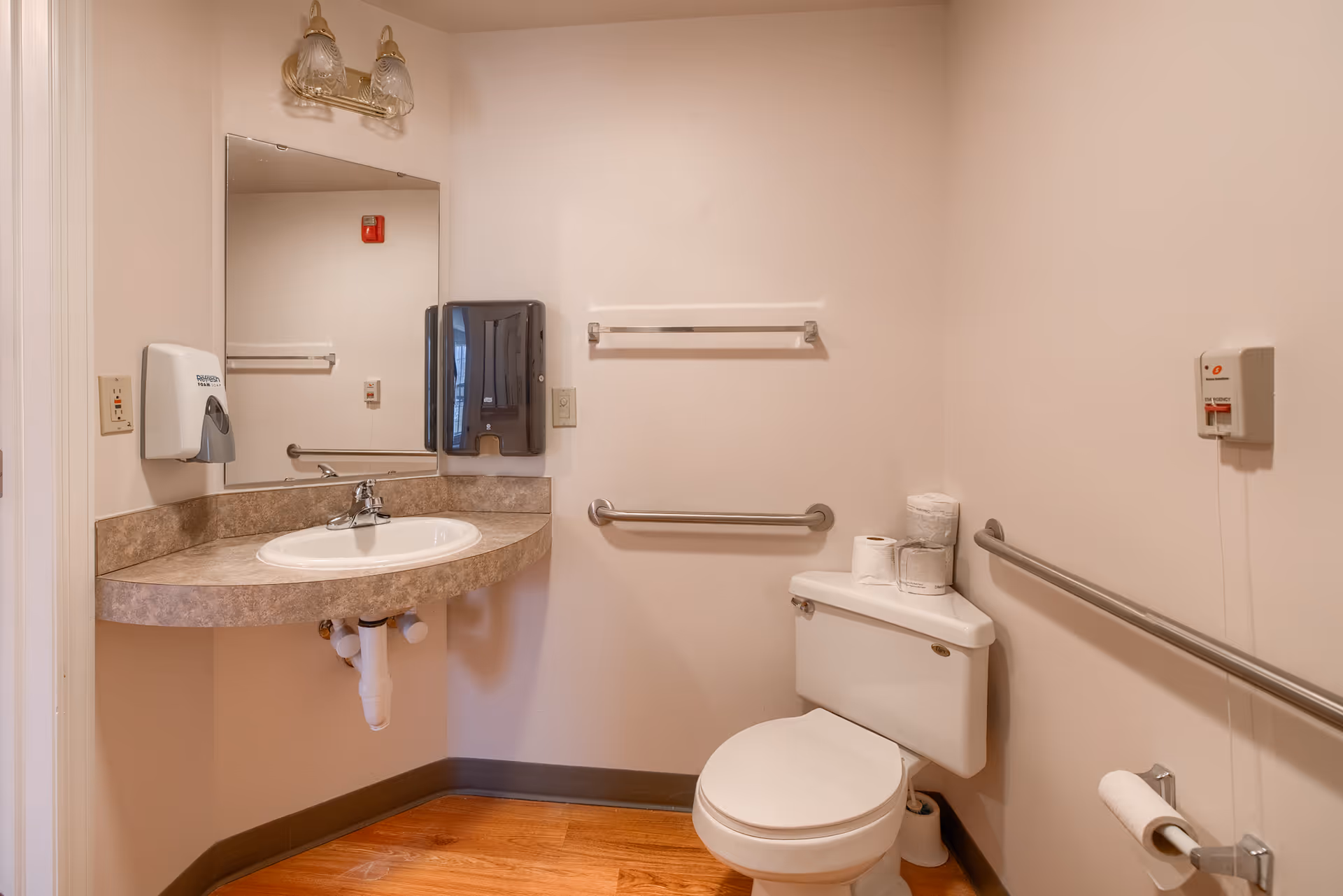 A clean and accessible bathroom featuring a wall-mounted sink with a large mirror above it, a soap dispenser, a paper towel dispenser, a toilet with several rolls of toilet paper on top, grab bars on the walls for support, and a wooden floor.