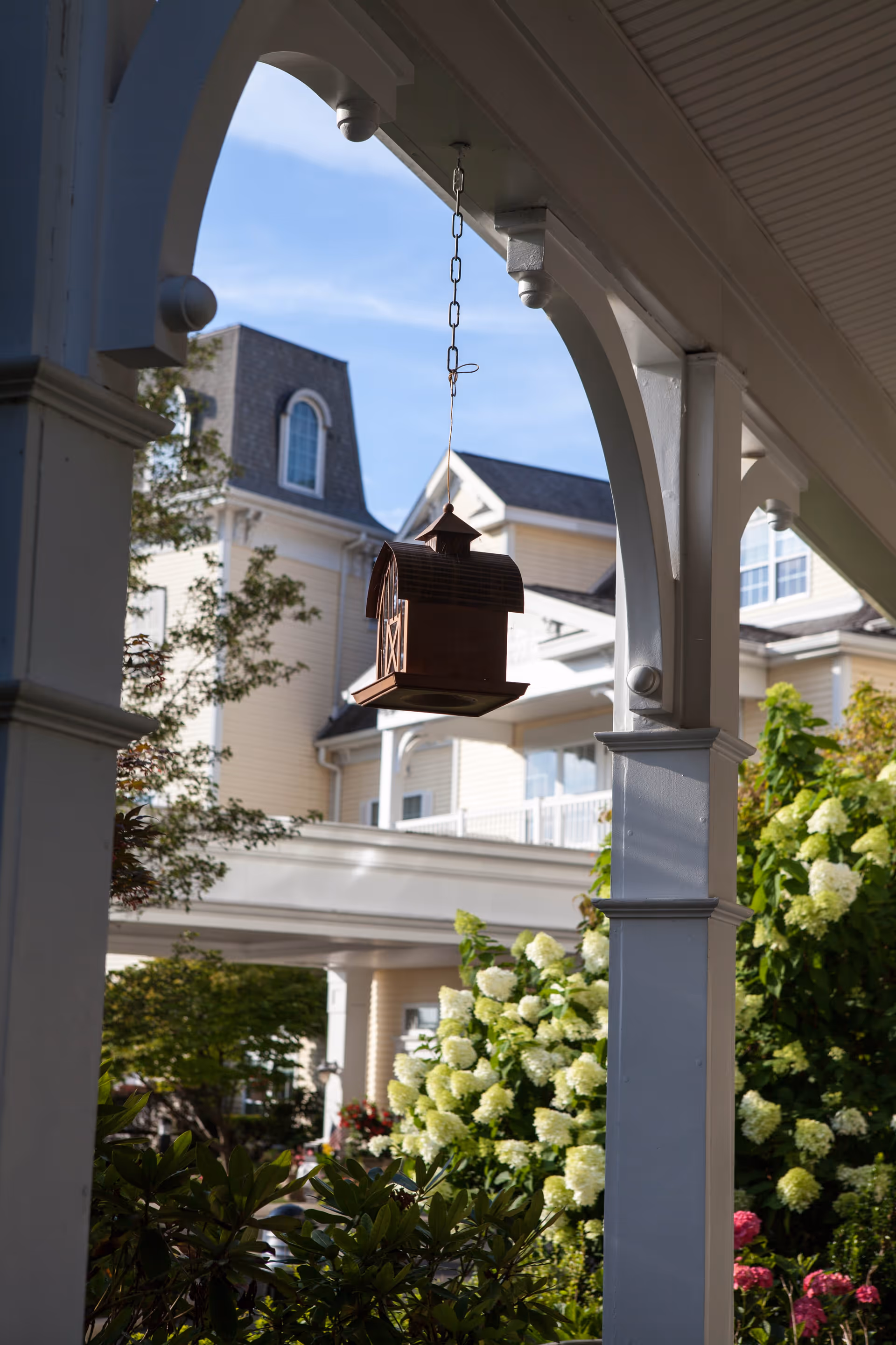 View of a hanging birdhouse shaped like a barn from a white porch with decorative arches, overlooking a garden with blooming white and pink flowers and a large beige building in the background under a blue sky.