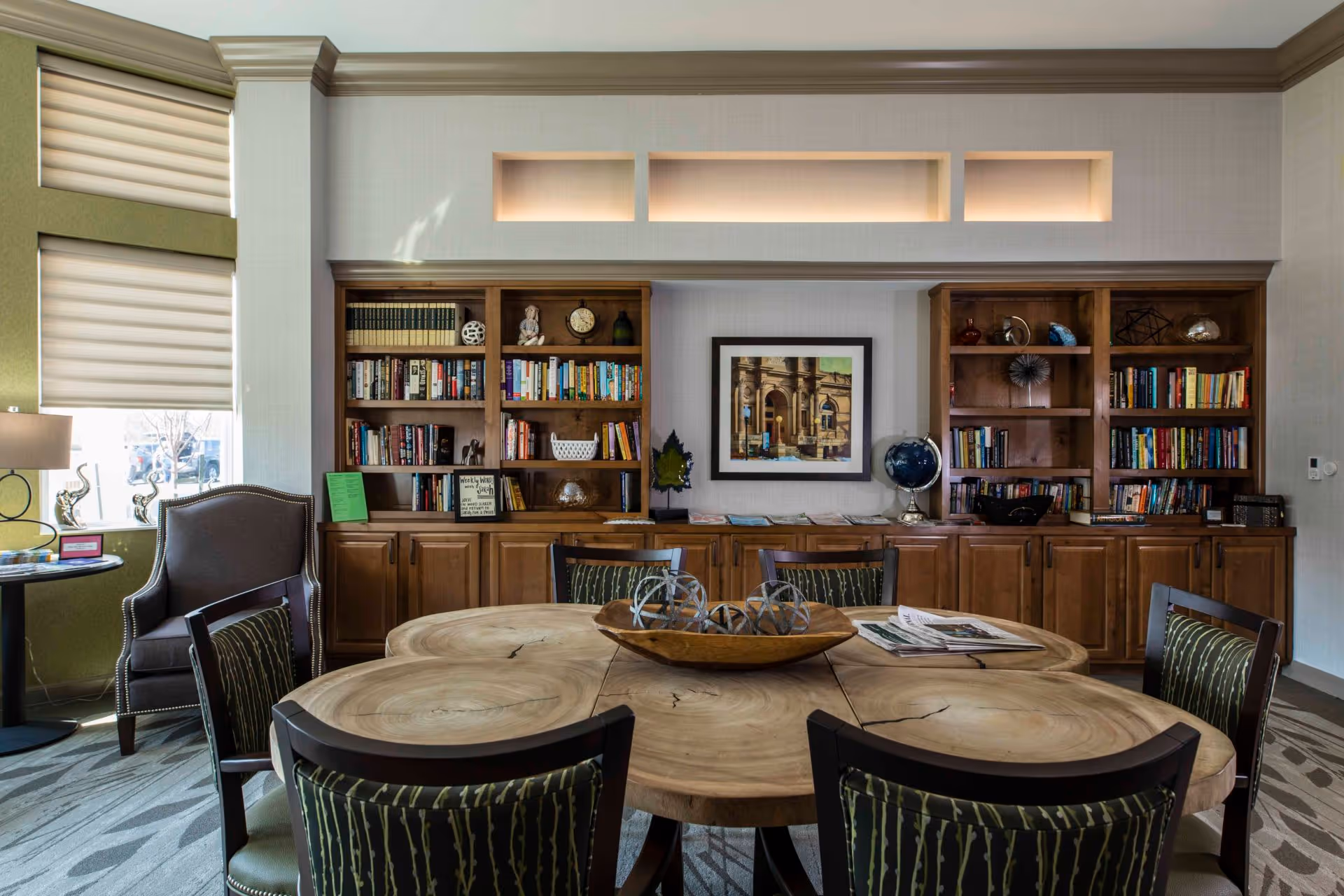 A cozy interior room with a large wooden table made of circular wood slices surrounded by six chairs with patterned upholstery. Behind the table is a built-in wooden bookshelf filled with books, decorative items, and a framed picture on the wall. A window with blinds and a lamp on a small round table are visible to the left.