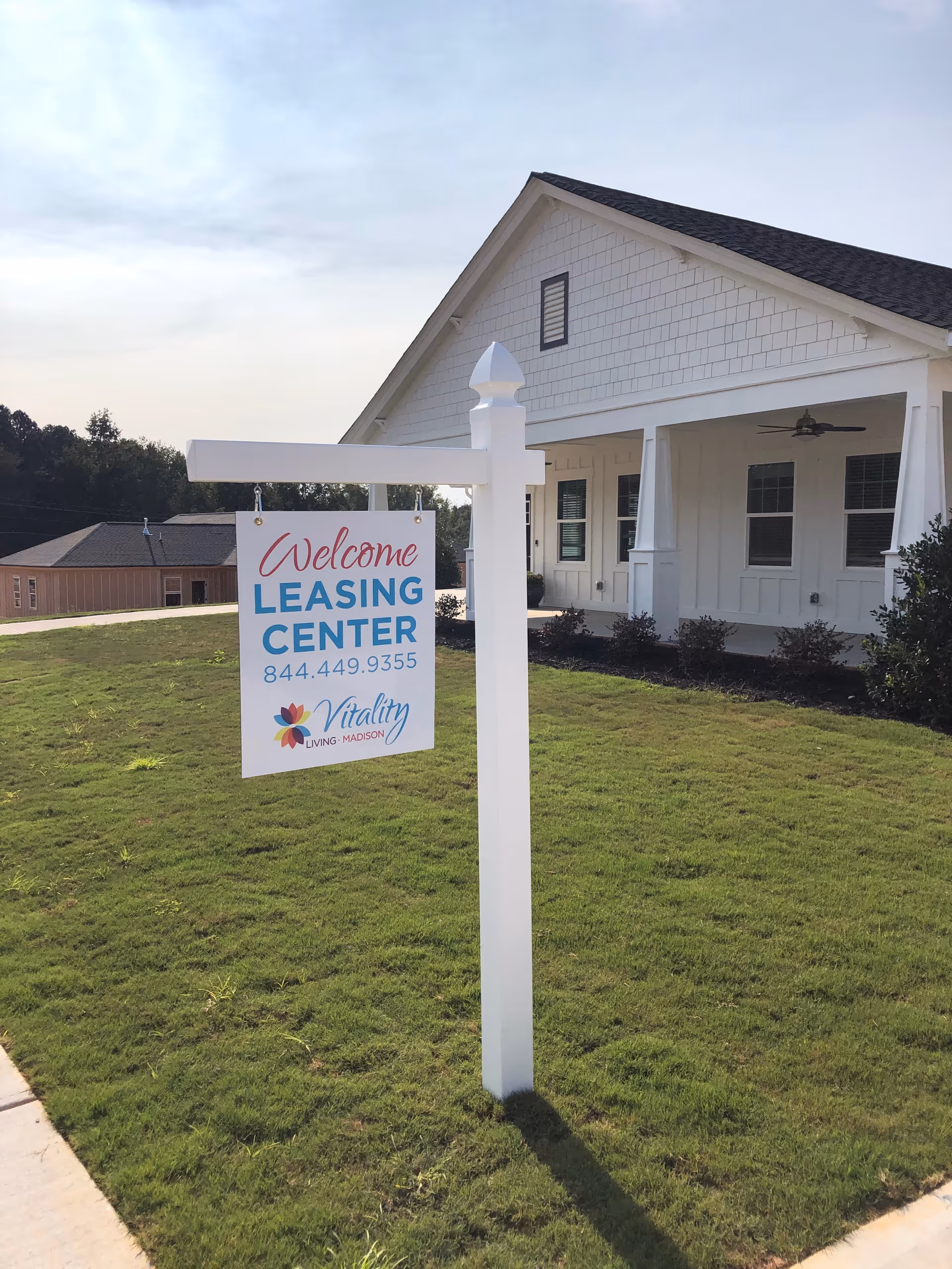 A white signpost on a grassy lawn with a sign that reads 'Welcome Leasing Center 844.449.9355 Vitality Living Madison' in front of a white building with a porch and ceiling fan.