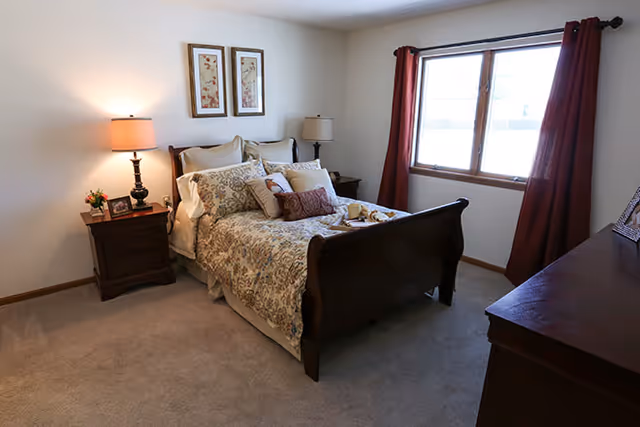 A cozy bedroom with a wooden bed frame and a floral patterned bedspread. The bed is adorned with multiple pillows and a tray with breakfast items. There are two bedside tables with lamps and a small flower arrangement on one. A large window with red curtains lets in natural light, and a wooden dresser is visible on the right side of the room. Two framed floral artworks hang above the bed.