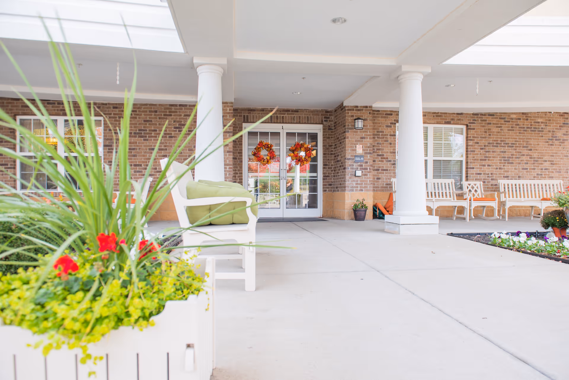 Entrance area of a senior living facility with white columns, brick walls, and glass double doors decorated with autumn wreaths. There are white benches with green cushions and planters with green plants and red flowers near the entrance.