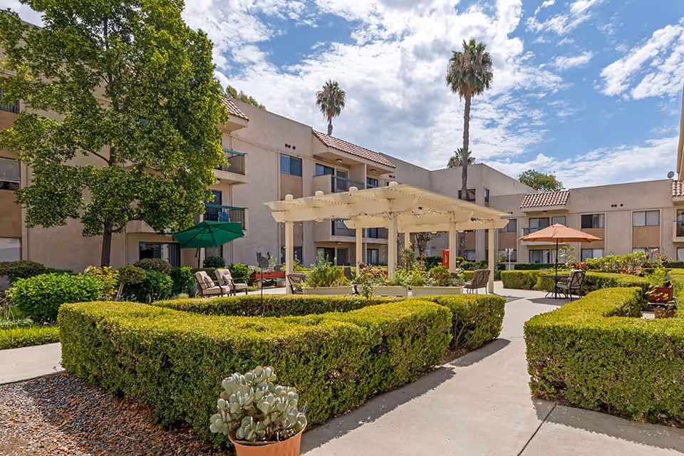 Outdoor courtyard area of a senior living facility with trimmed hedges, a white pergola, patio chairs, tables with umbrellas, and a large tree. The building with balconies and windows surrounds the courtyard under a partly cloudy sky.
