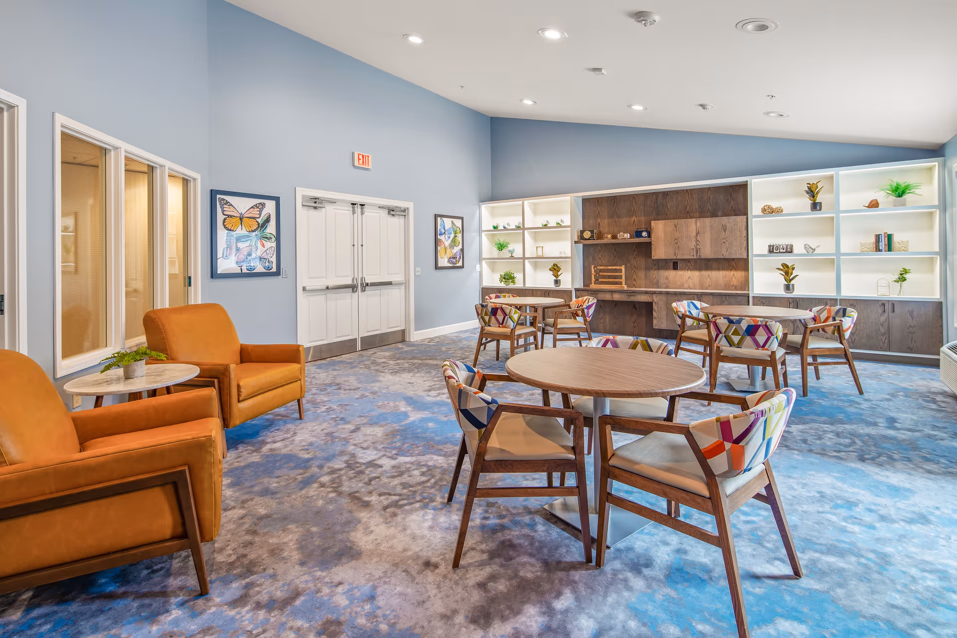 A bright and spacious common area with blue walls and carpet featuring a cloud-like pattern. The room contains several round wooden tables surrounded by colorful patterned chairs. There are two orange armchairs with a small round side table between them on the left side. The back wall has built-in shelves with decorative plants and items, and there are two framed butterfly artworks on the wall near double doors.