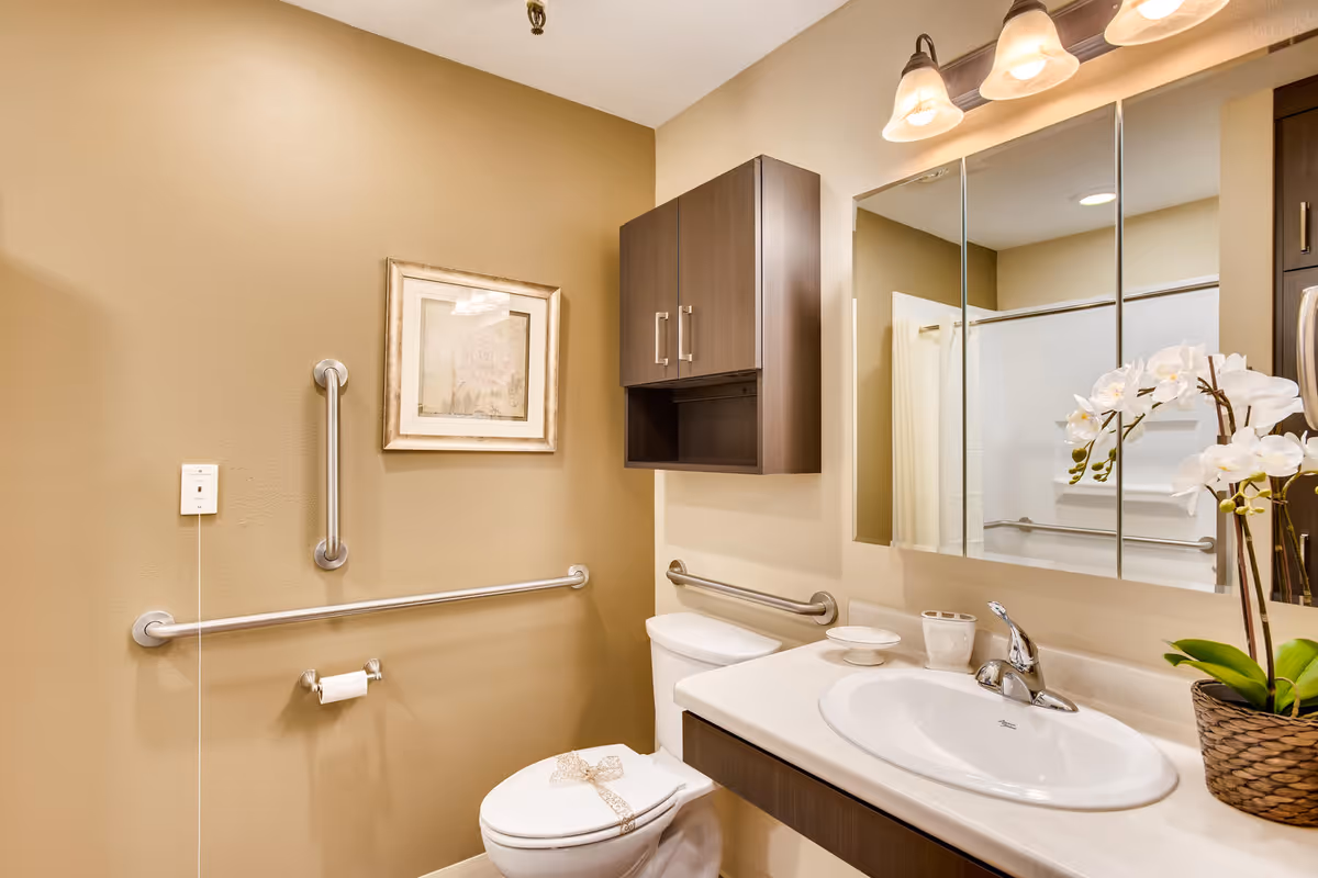A clean and well-lit bathroom with beige walls, a white toilet with a decorative bow on the seat, a white sink with a chrome faucet, a large mirror above the sink, a wooden cabinet mounted on the wall, grab bars for accessibility, a framed floral picture on the wall, and a potted orchid plant on the countertop.