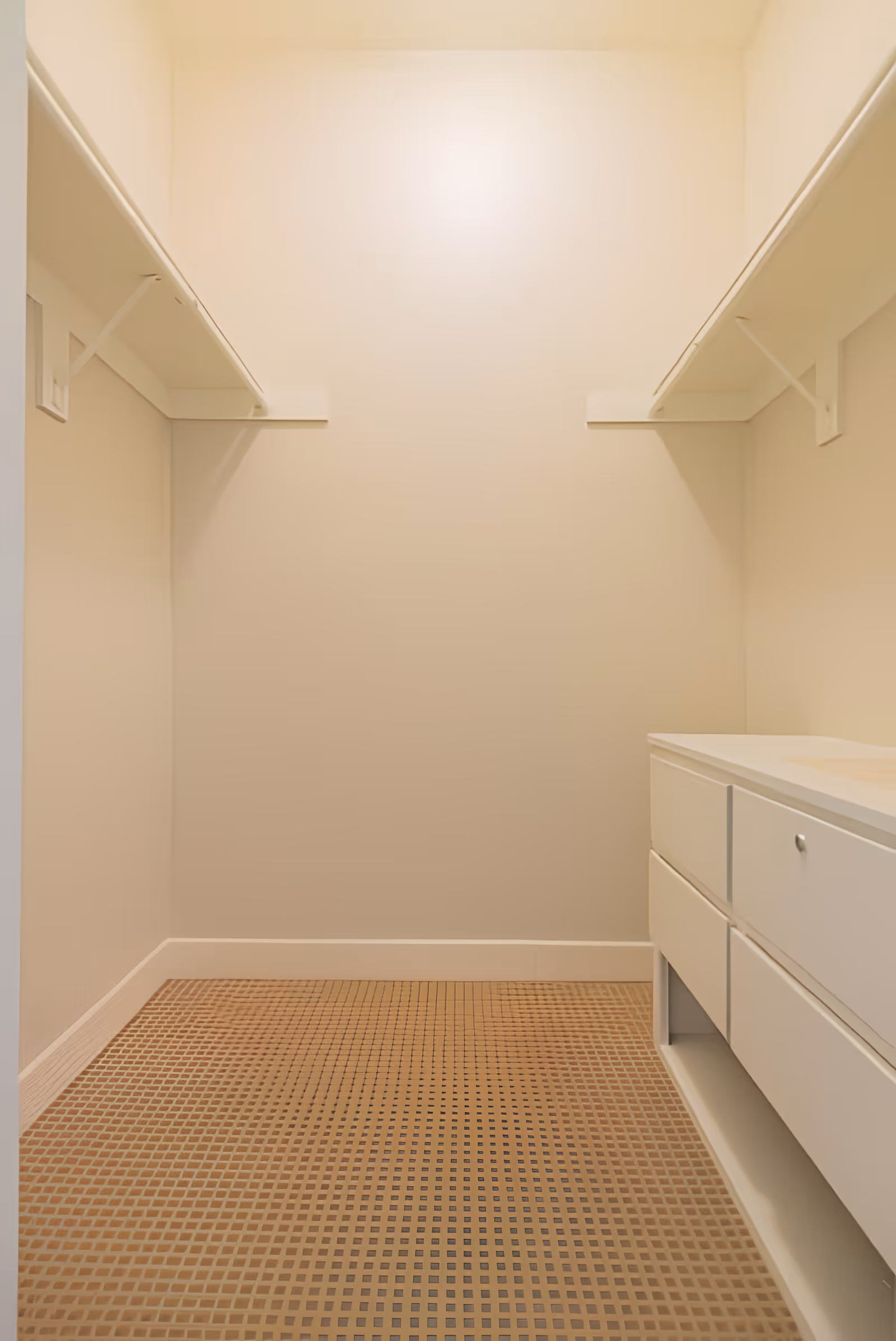 Empty walk-in closet with beige walls, two white shelves on either side near the ceiling, and a white cabinet with drawers on the right side. The floor has a brown checkered pattern.