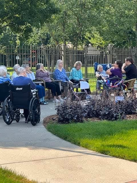 A group of elderly people sitting outdoors in wheelchairs and chairs along a paved pathway near a garden with shrubs and grass. They are gathered in a shaded area with trees and a metal fence in the background, enjoying the outdoor environment.