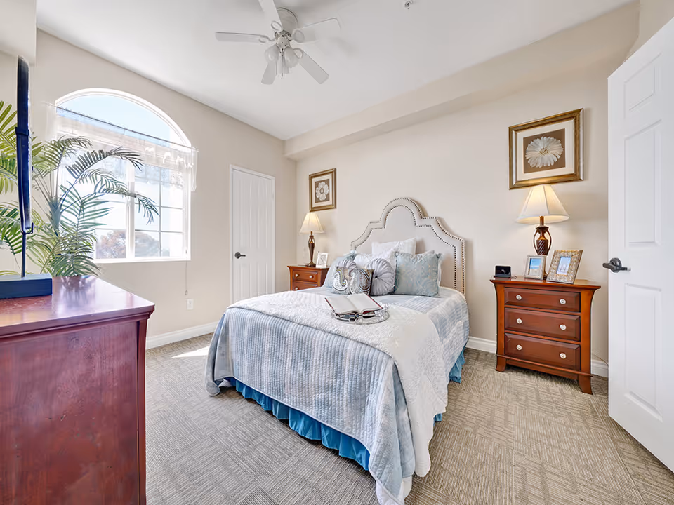A bright and cozy bedroom with a neatly made bed featuring multiple pillows and a light blue quilt. The room has beige walls, a ceiling fan, two wooden nightstands with lamps and picture frames, a wooden dresser with a plant on top, and a large arched window letting in natural light.