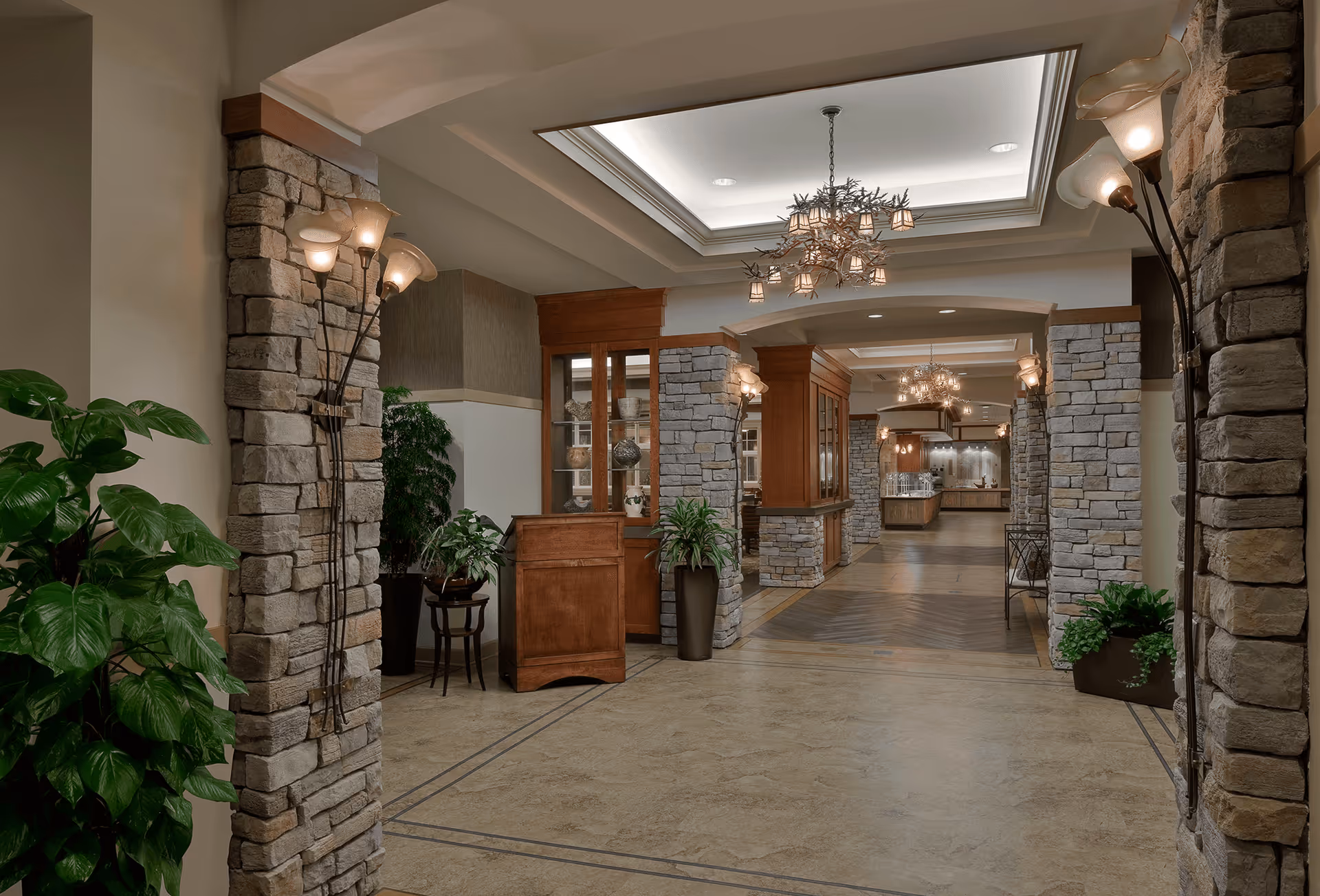 Well-lit stone-columned lobby with chandeliers, potted plants, a wooden reception desk, and a view toward a dining/kitchen area.