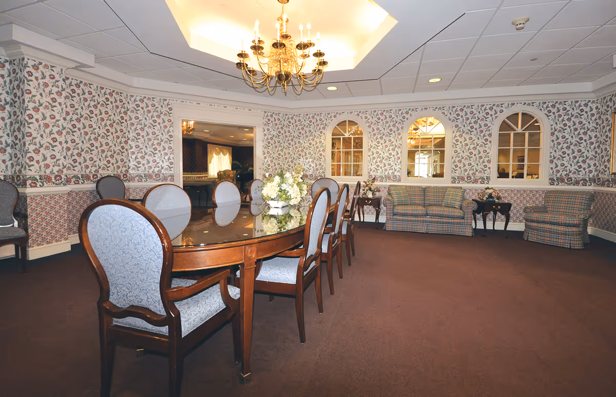 A formal dining room with a long wooden table surrounded by upholstered chairs. The walls are decorated with floral wallpaper and three arched mirrors. There is a chandelier hanging from the ceiling, and a seating area with a plaid sofa and armchair is visible in the background.