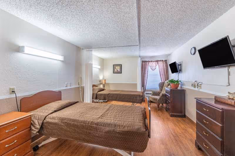Nursing facility bedroom with an adjustable single bed covered in brown bedding, wooden dressers, a wall-mounted TV, an armchair, and a mirrored wall.