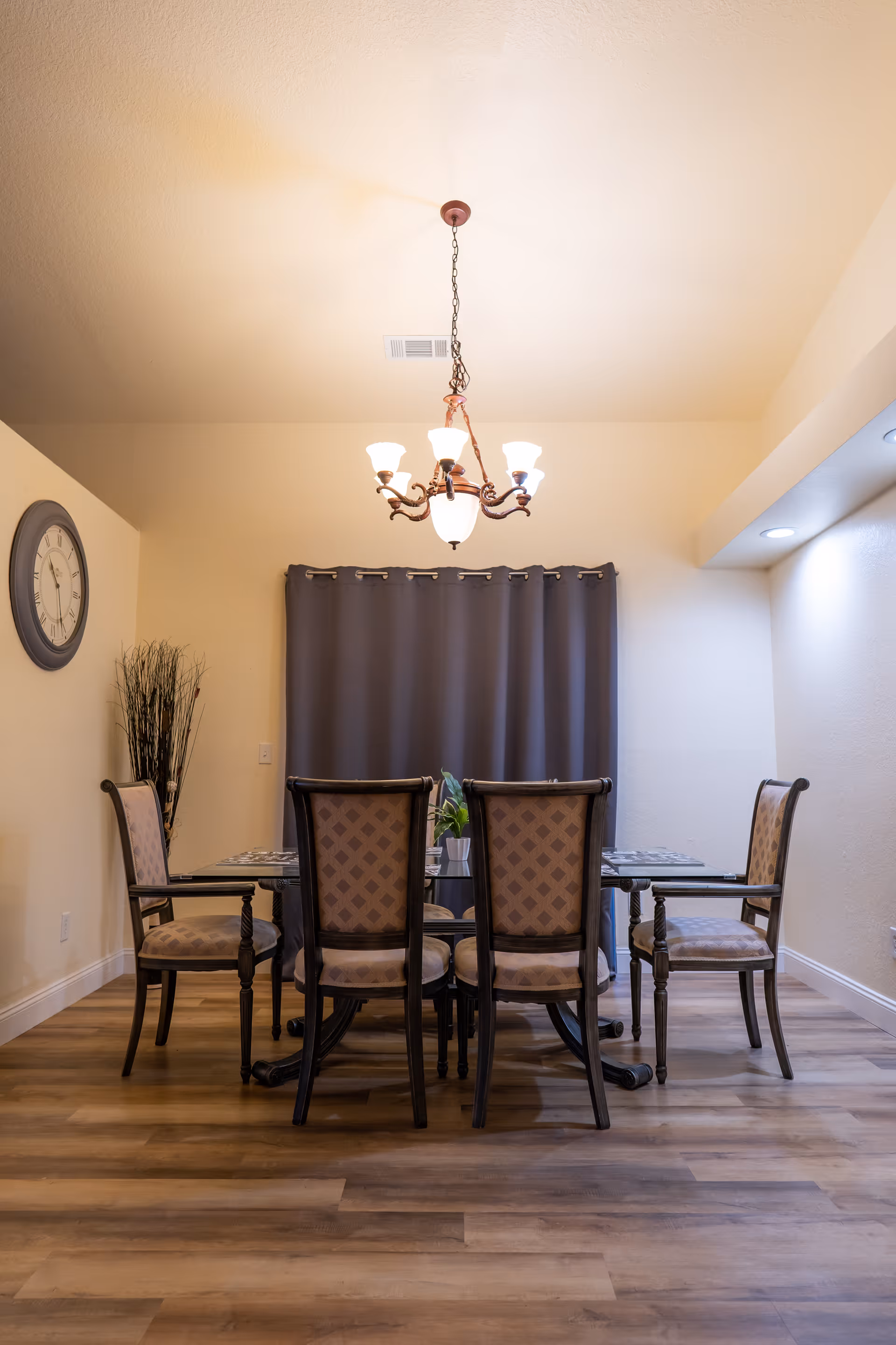 Dining room with a rectangular table and six upholstered chairs under a chandelier, gray curtains and a wall clock on a wood floor.