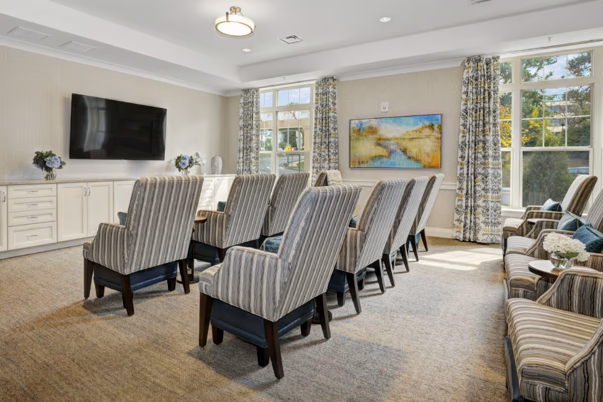 Bright common seating room with rows of striped armchairs facing a wall-mounted TV and large windows.