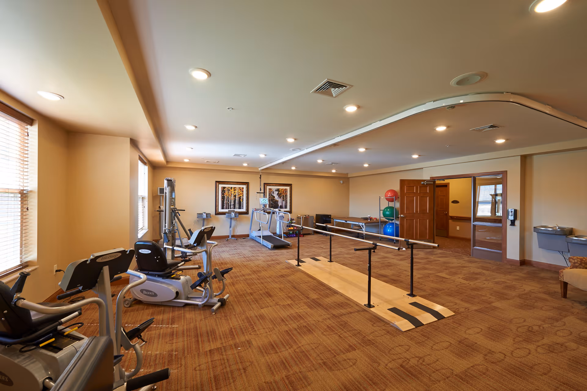 A spacious senior living fitness room with exercise equipment including stationary bikes, a treadmill, and parallel bars for walking support. The room has beige walls, carpeted floor, recessed lighting, and windows with blinds. There are colorful exercise balls stacked on a rack and framed artwork on the far wall.