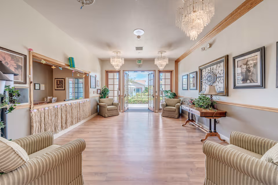 A bright and spacious interior hallway of a senior living facility with wooden flooring, four striped armchairs, a wooden console table with a lamp and plant, framed pictures on the walls, and three elegant chandeliers hanging from the ceiling. Double glass doors at the end open to an outdoor garden area.