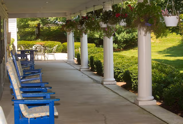 Covered outdoor patio area with blue wooden rocking chairs lined up along one side and white columns supporting the roof. Hanging flower baskets with colorful blooms are suspended from the ceiling. In the background, there is a small table with metal chairs and green bushes and trees surrounding the area.