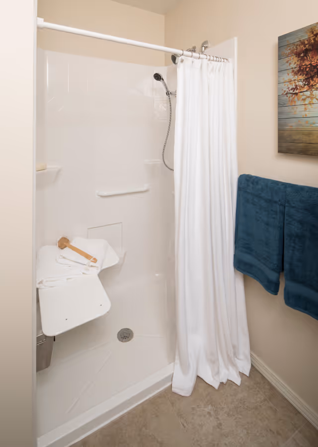 A clean, accessible shower area with a white fold-down seat holding folded white towels and a wooden bath brush. The shower has a white curtain, a handheld showerhead, and a grab bar on the wall. Two dark blue towels hang on a rack on the adjacent wall, and a decorative painting with autumn leaves is visible above the towels.