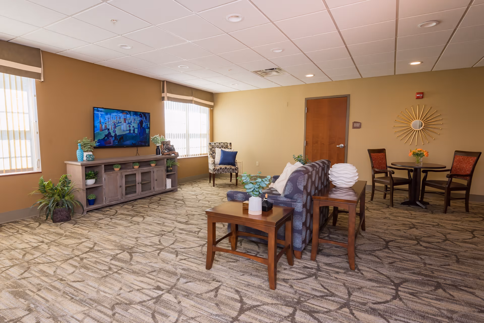 A well-lit senior living facility common area with a patterned carpet, a couch with pillows, a wooden TV stand with plants and decorations, a wall-mounted flat-screen TV, a single armchair with a blue pillow, and a small round table with two chairs and a flower vase. The walls are painted beige, and there is a decorative sunburst mirror on the wall near a wooden door.